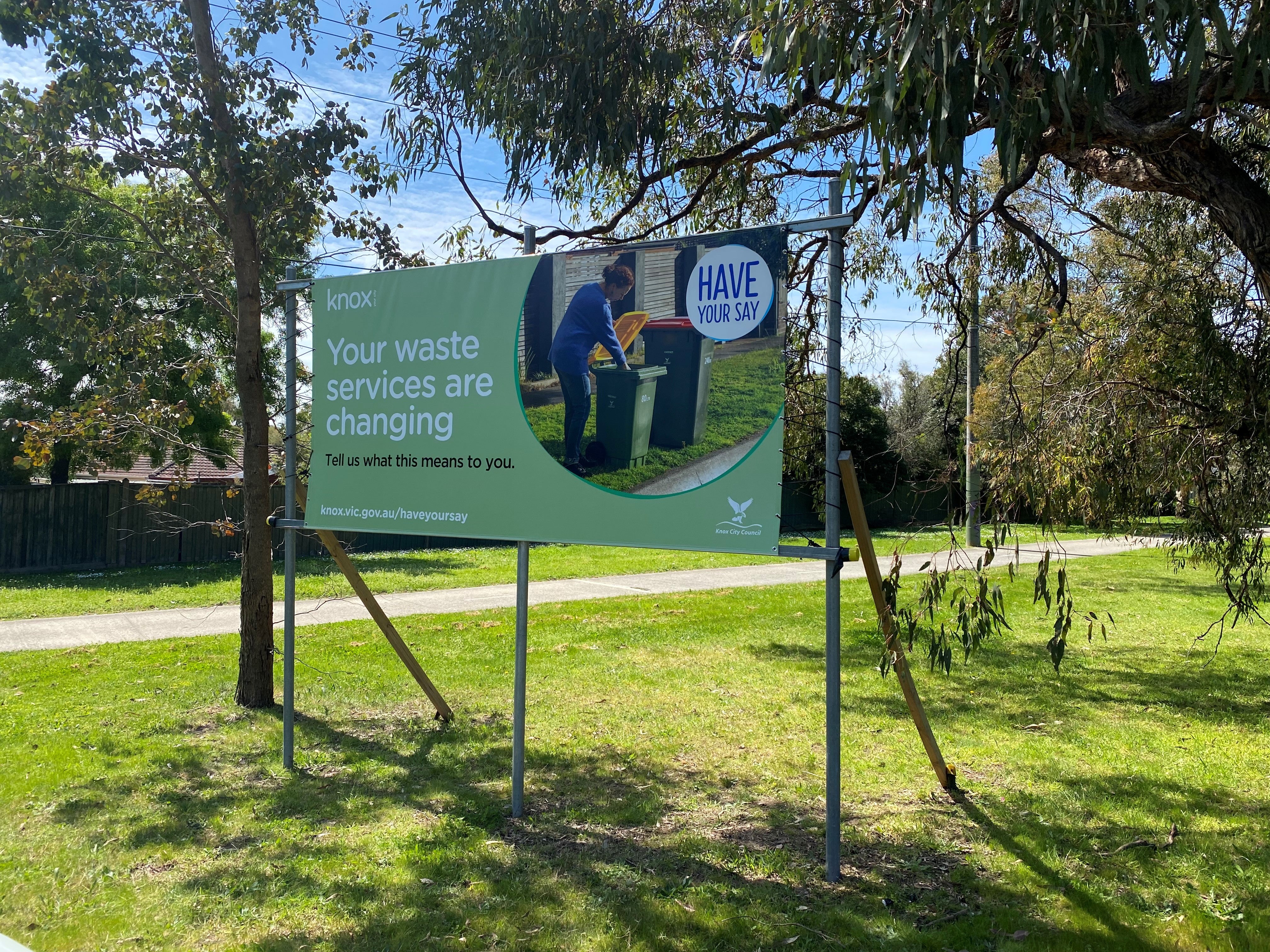 A large outdoor banner sign on metal posts in a grassy park area. The sign has a light green background with white text reading 'Your waste services are changing' and a circular photo of a person disposing of trash.